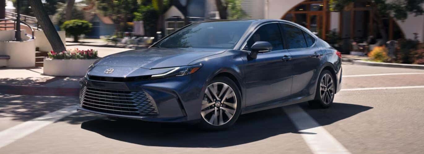 Exterior three-quarter view of a blue Toyota Camry making a turn on a sunny city campus road.