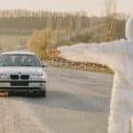 Photo of a man in a hazmat suit stopping a white BMW with a person wearing a mask.