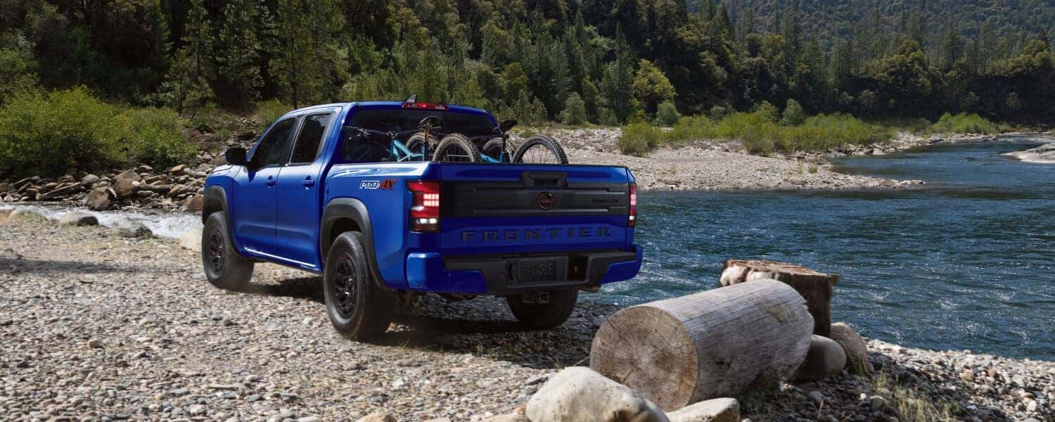 A rear image of blue Nissan Frontier with bikes in its truck bed, parked next to a river by a forest
