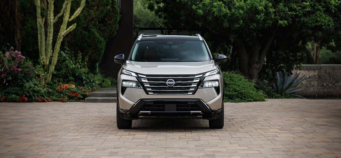 A front view of a silver Nissan Rogue parked on a tiled ground near a garden