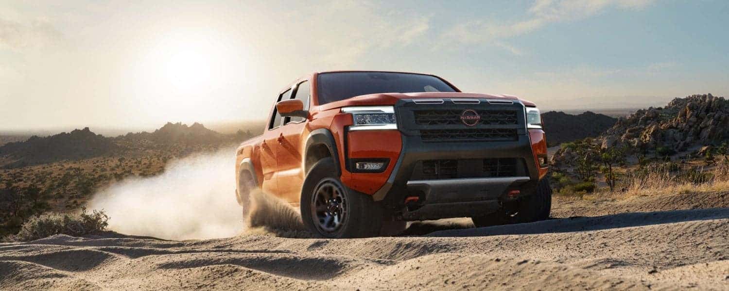 An orange Nissan Frontier leaving behind a dust trail while it drives over a dirt road in the desert