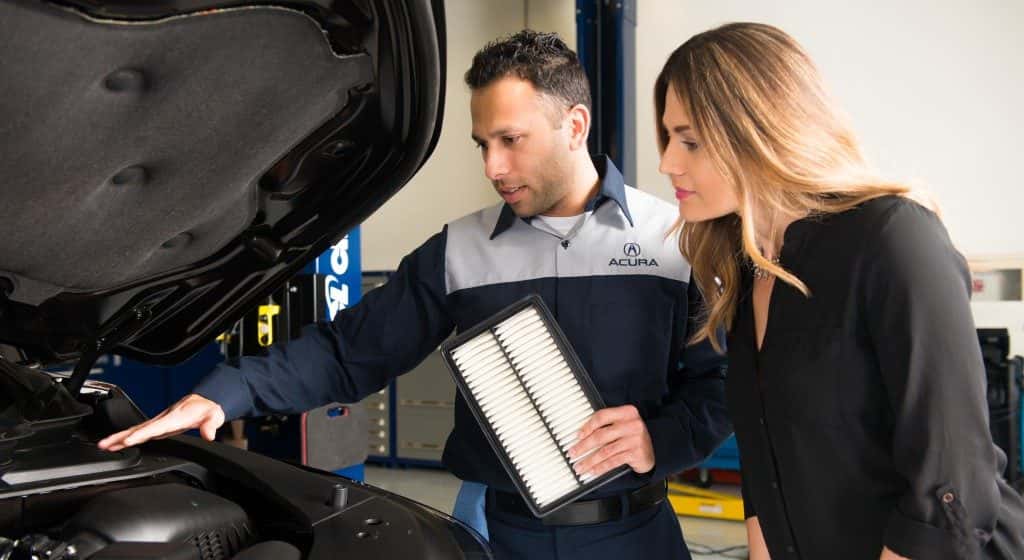 An Acura technician holding an engine air filter and showing it to a customer beside an open hood