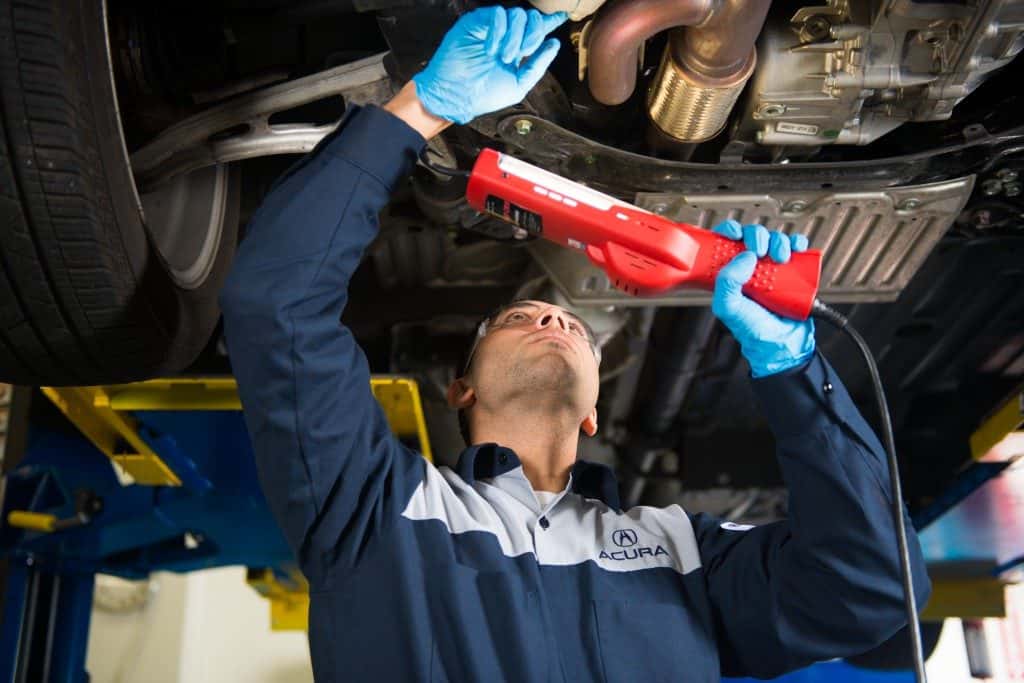 An Acura technician using a handheld inspection lamp to examine the underside of a raised vehicle, with the front wheel and subframe in view