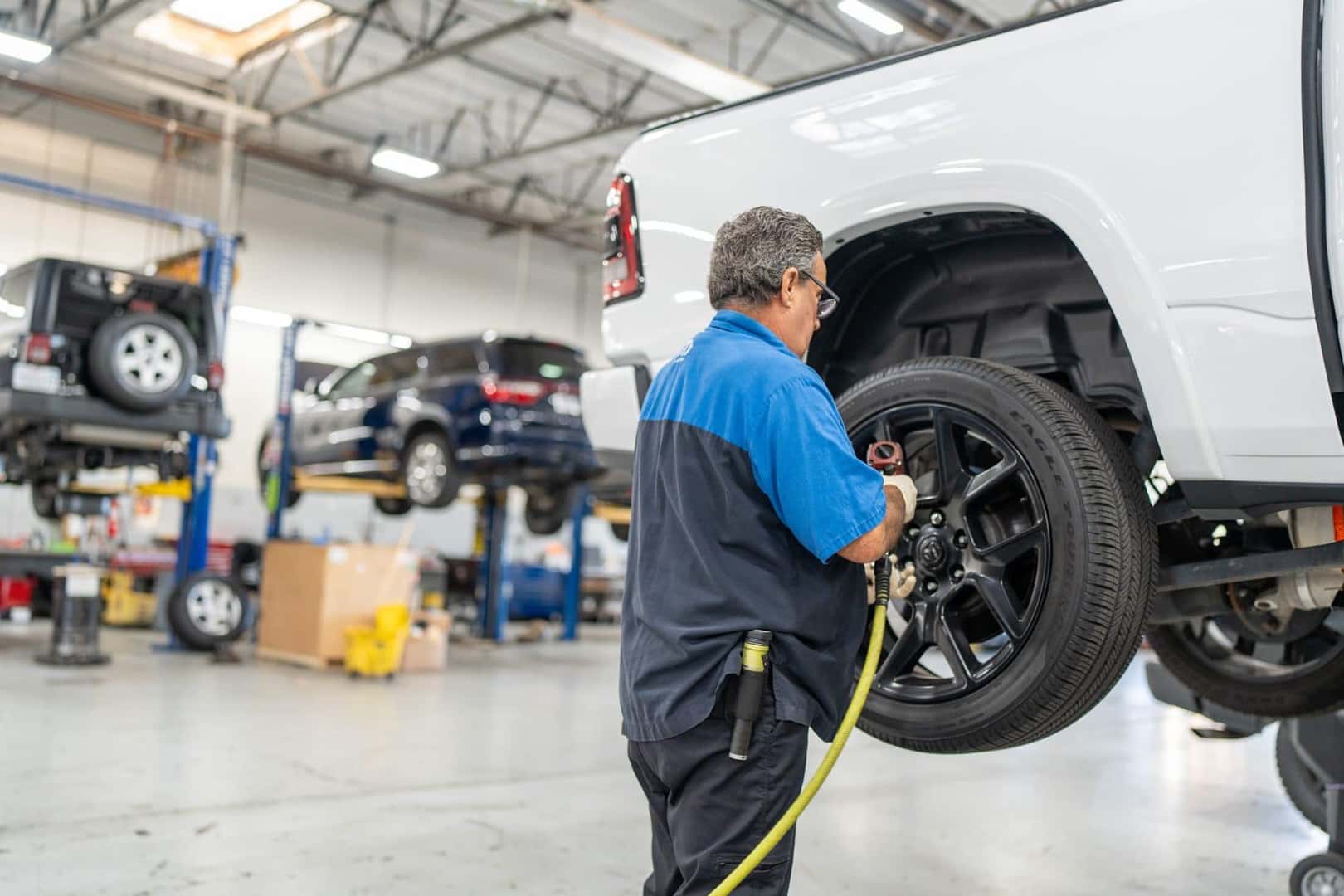 A technician using an air impact wrench on the wheel of a white Ram 1500 raised on a lift in the Elk Grove Dodge Chrysler Jeep Ram service shop