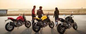 Three Ducati Monster motorcycles lined up while riders stand nearby overlooking the beach