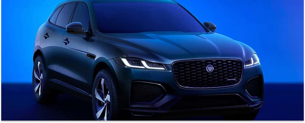 A dark green 2026 Jaguar F-Pace SUV with a prominent black mesh front grille and sleek LED headlights is showcased in a studio setting against a vibrant blue background.