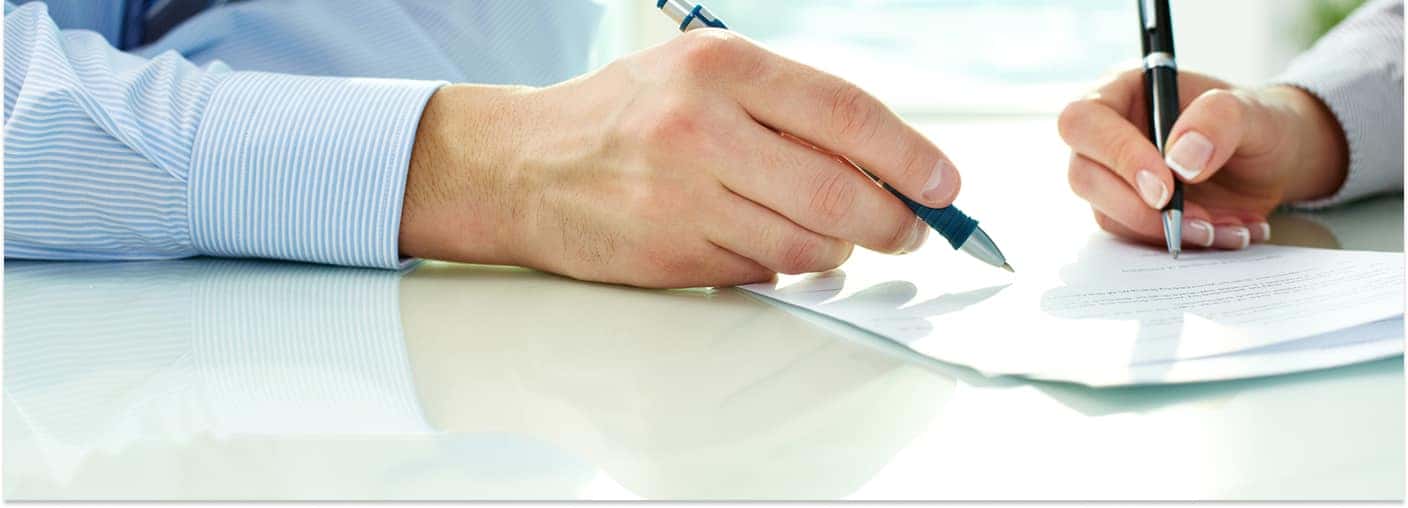 A close-up view of two people holding pens as they sign or review paperwork on a bright, reflective desk.