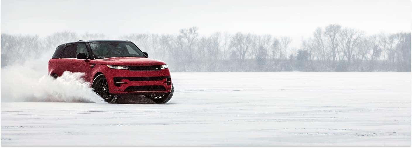 A front three-quarter view of a red 2025 Range Rover Sport driving dynamically across a snow-covered landscape and kicking up a large plume of snow.