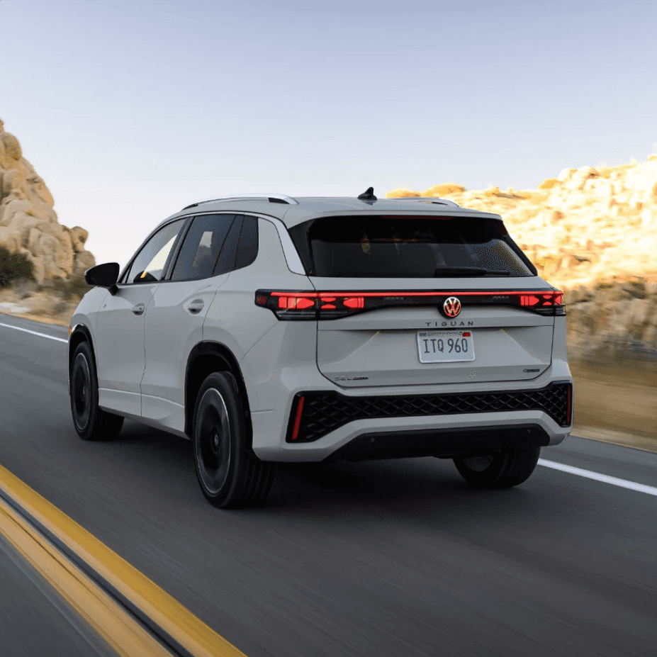 Rear exterior view of a 2026 Volkswagen Tiguan driving on a scenic open road, showcasing LED taillights and modern SUV design