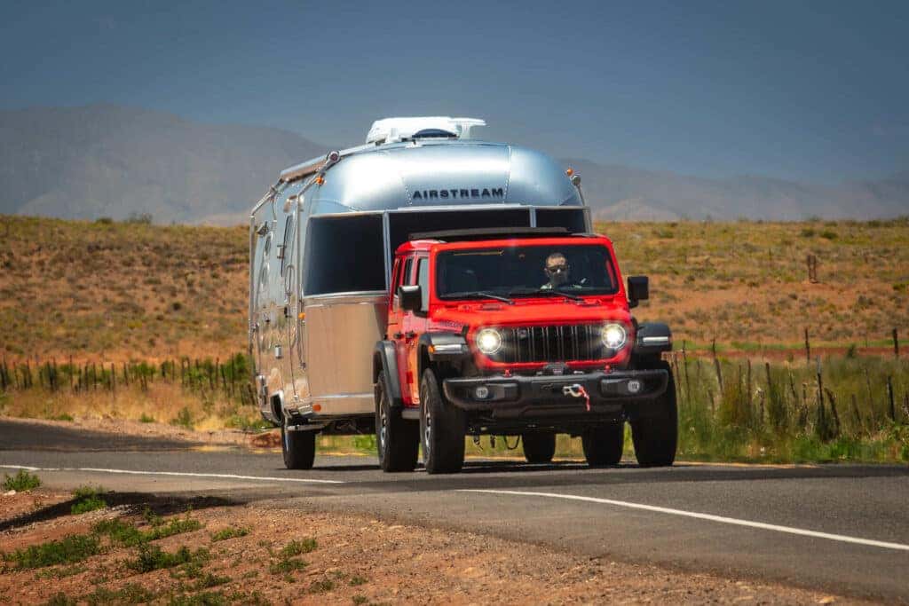 A red Jeep Wrangler pulling a silver metallic camper behind it.