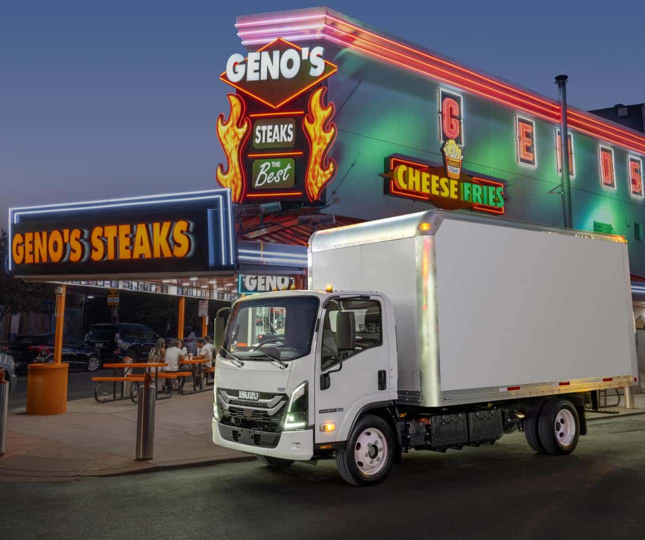 Isuzu NRR EV parked outside a neon-lit restaurant setting at dusk.