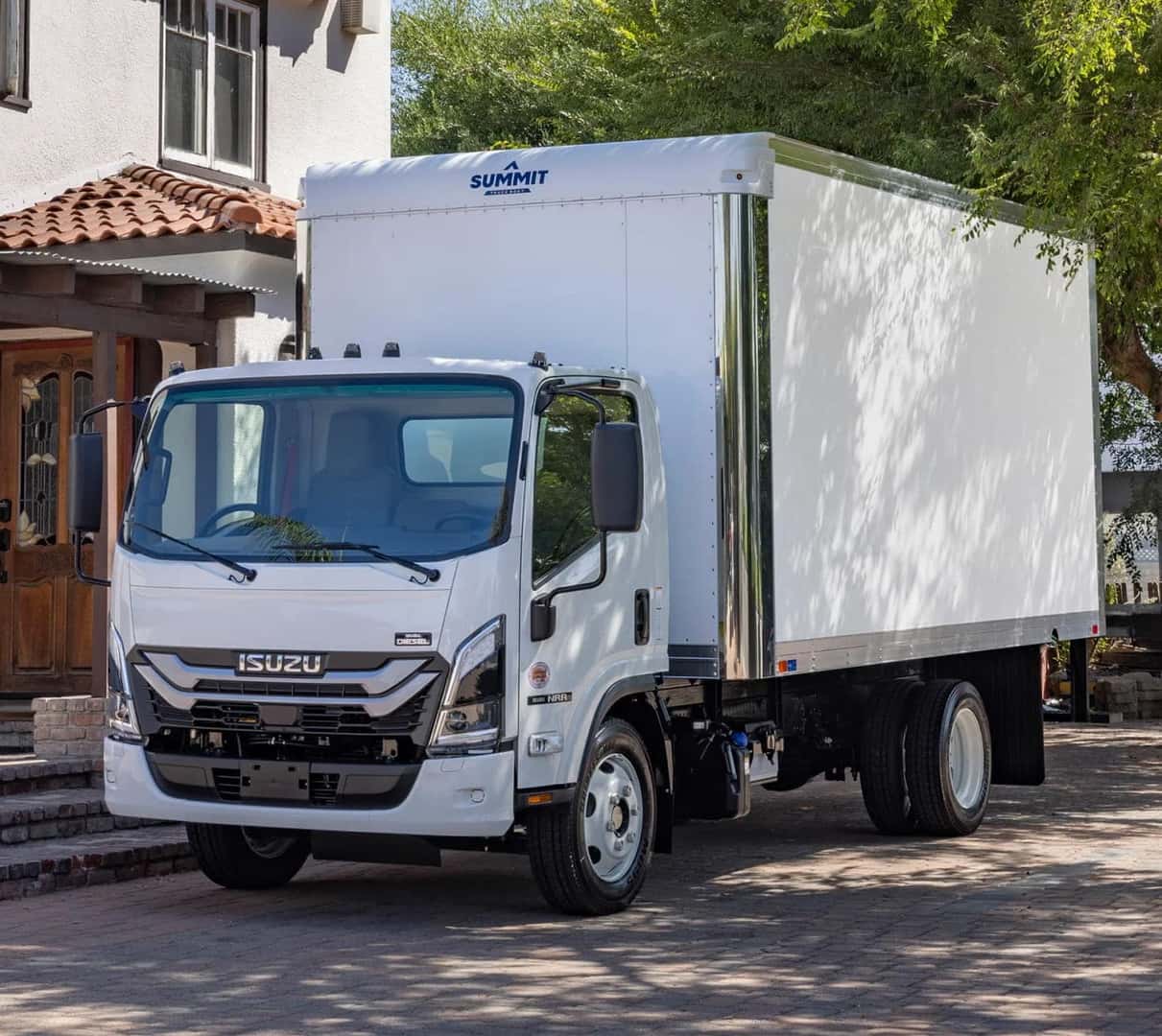 A white Isuzu box truck is parked on a sunny residential street with trees and a house in the background.