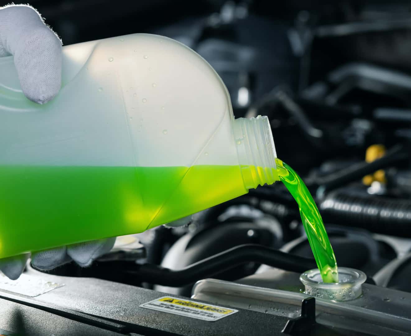 A close-up of a gloved hand pouring green coolant into an open radiator.