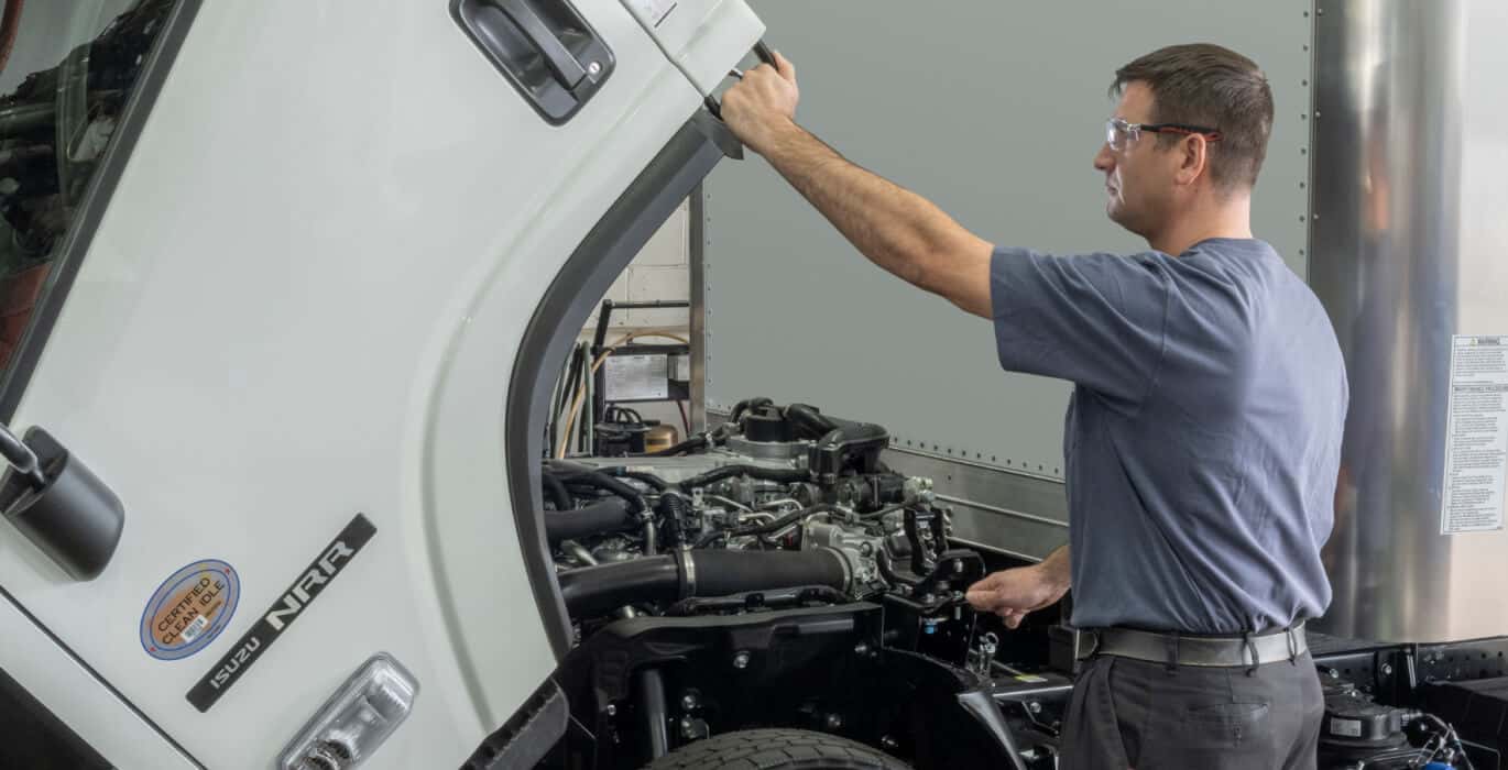 A man in safety glasses lifts the cab of an Isuzu N-Series exposing mechanical components underneath.