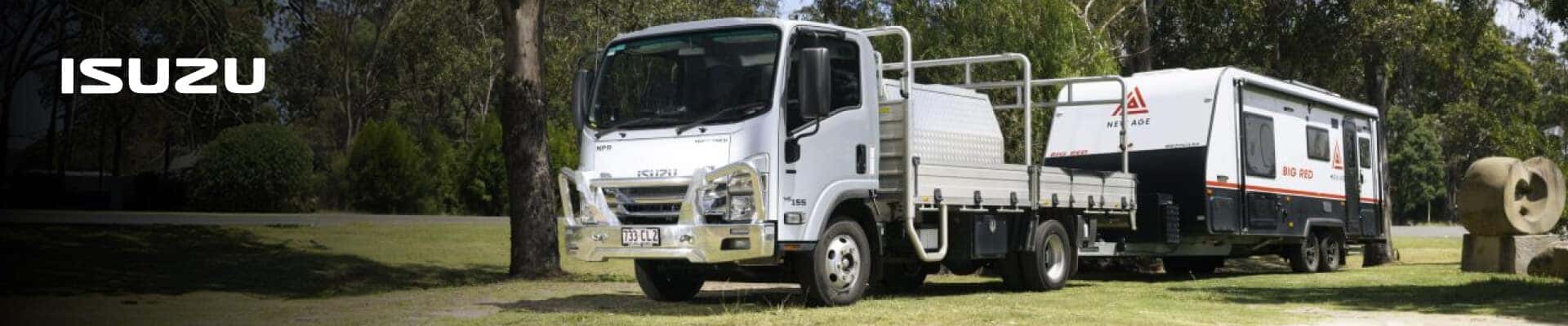 An Isuzu NPR flatbed towing a large trailer in a sunny wooded park.