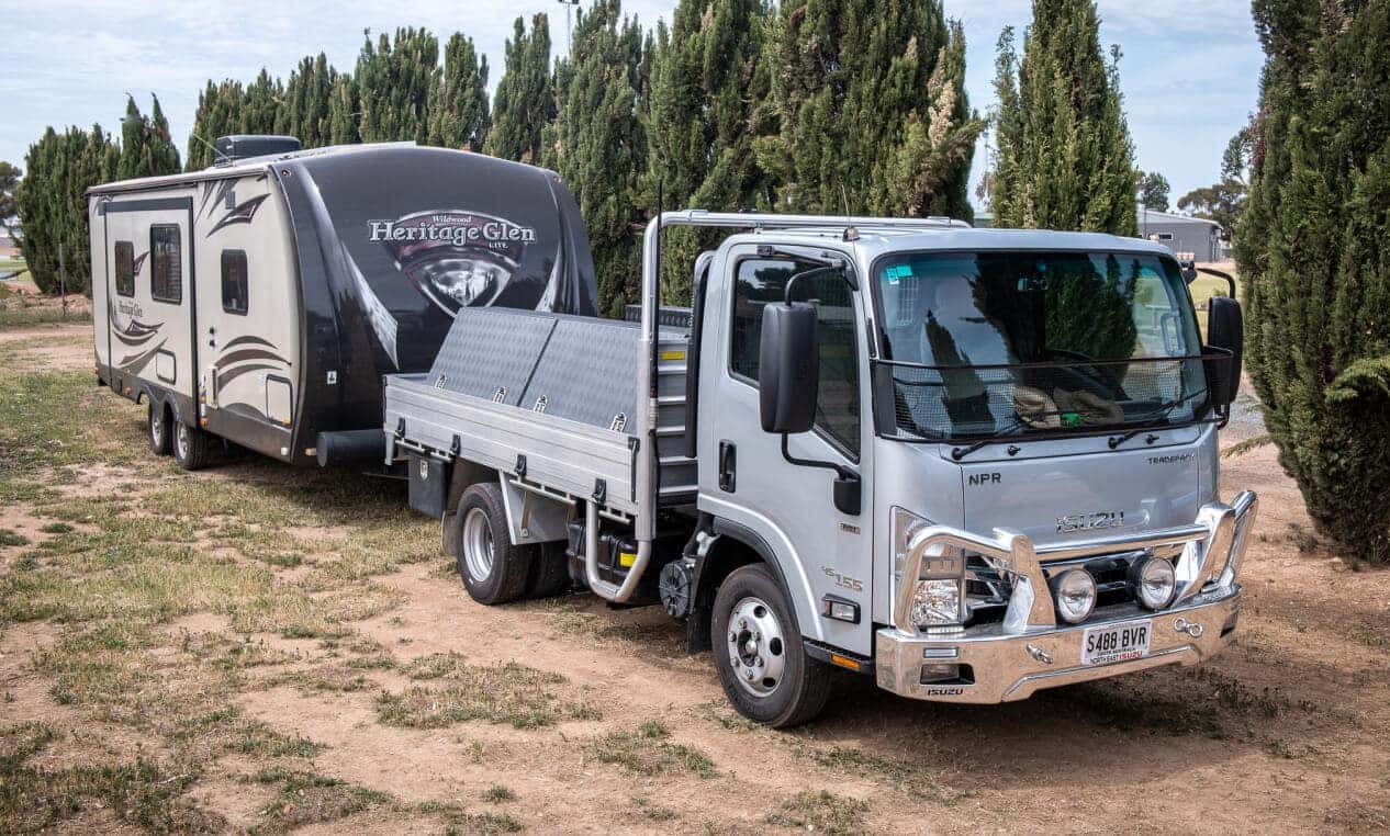 An Isuzu NPR flatbed towing a large camper trailer along a tree-lined dirt road.