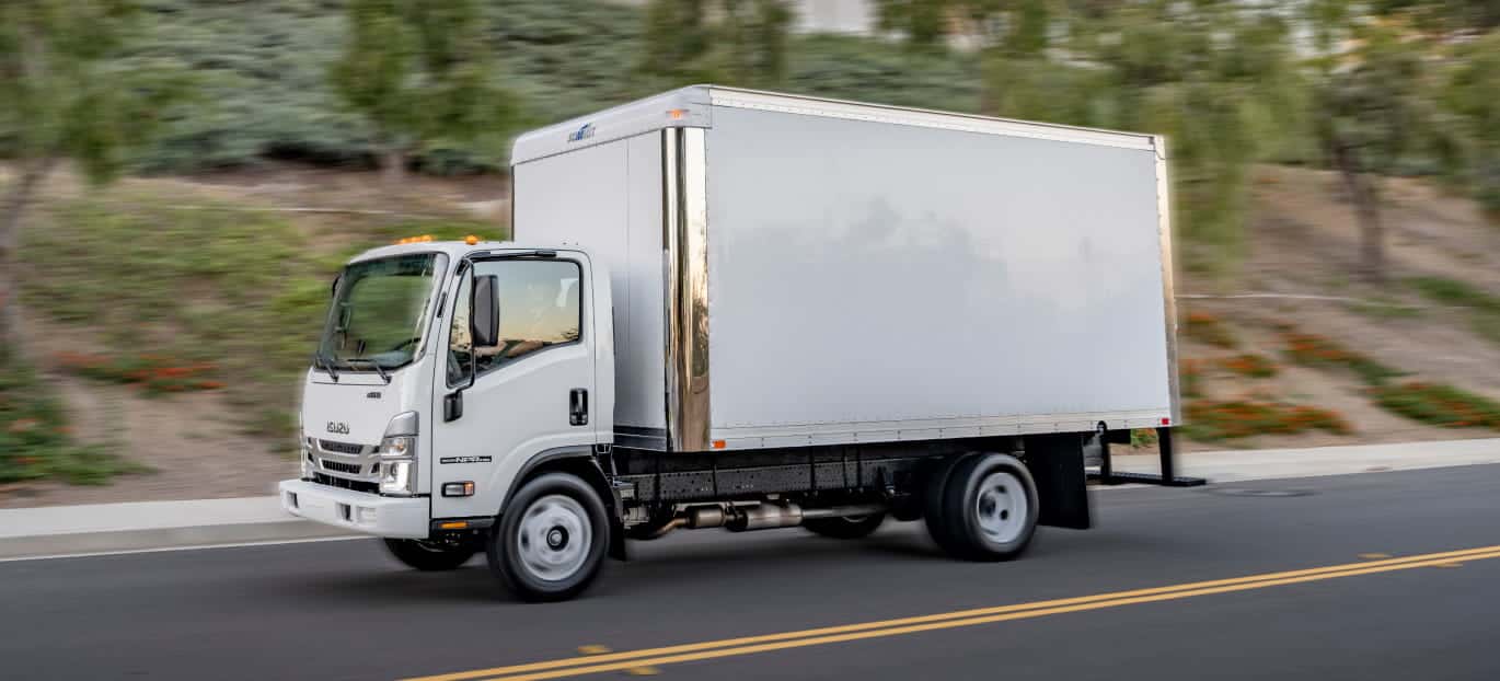 White Isuzu box truck driving on a paved road with trees in the background.
