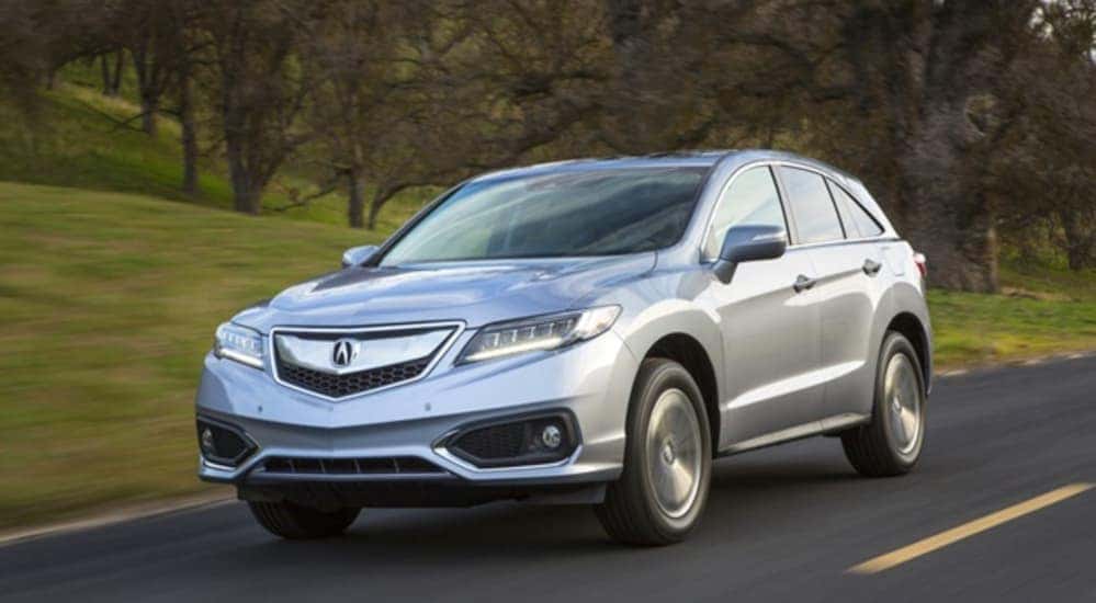 A silver 2016 Acura RDX is driving on a tree lined road.