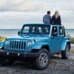 A couple are sitting on their 2018 Jeep Wrangler, which they found while searching "used car dealerships near me", while overlooking a large lake off a dirt trail.