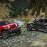 A red 2-door and a grey 4-door 2020 Jeep Wrangler are parked next to a small stream in the woods.