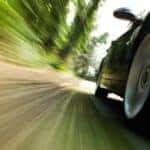 A close up of a muscle car driving past blurred trees is shown.