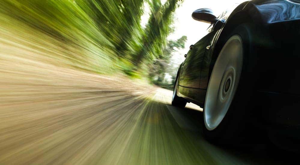 A close up of a muscle car driving past blurred trees is shown.
