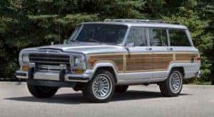 A blue and wood-paneled 1989 Jeep Grand Wagoneer is parked in front of trees.