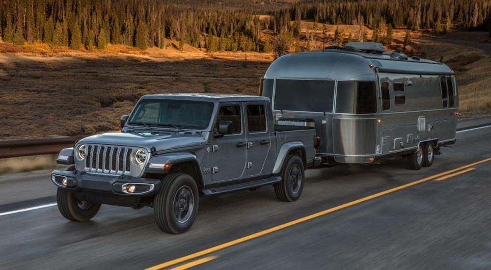 A silver 2020 Jeep Gladiator is towing a silver camper on a highway after leaving a Costa Mesa Jeep dealership.