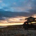 The silhouette of a Jeep Wrangler with a roof tent is shown against a cloudy sunset.