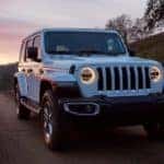 A white 2020 Jeep Wrangler Unlimited is shown from the front driving on a rural highway at sunset.
