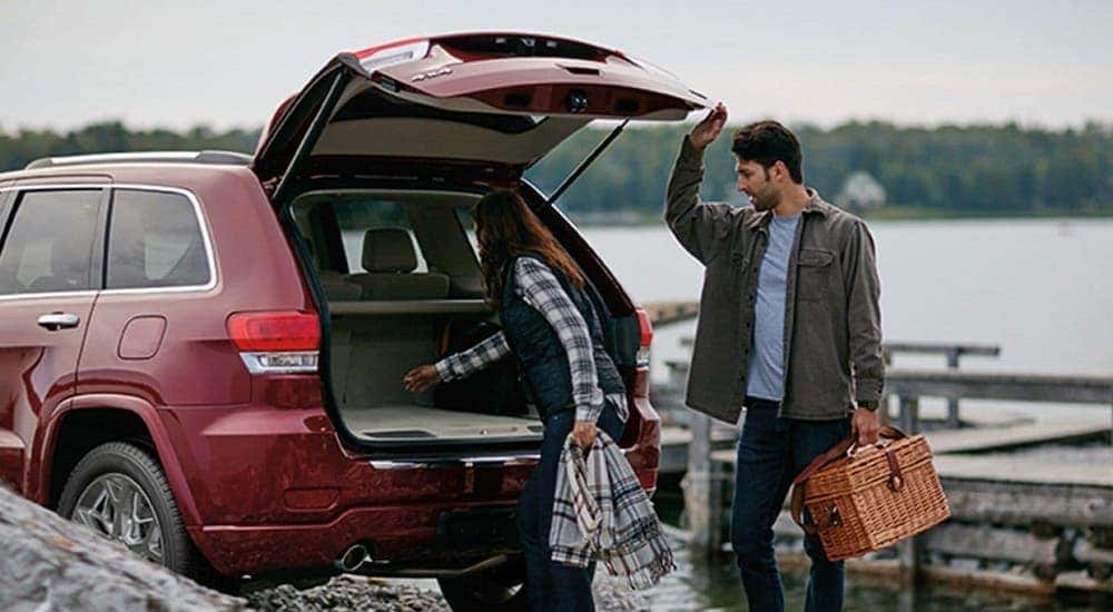 A couple is unloading cargo from the rear of a red 2020 Jeep Grand Cherokee with a lake in the background.