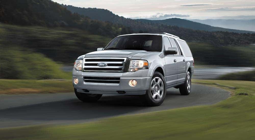 One of the best used SUVs, a silver 2013 Ford Expedition, is driving on a road past green hills.