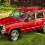 A red late 1980s Jeep Cherokee XJ is parked on grass in front of a cabin.
