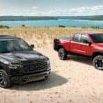 Two Ram trucks for sale, a black and a red 2020 Ram 1500, are parked on sand in front of an ocean bay.
