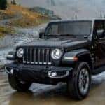A black 2018 used Jeep Wrangler Unlimited is shown driving alongside a river on a dirt road in the rain.