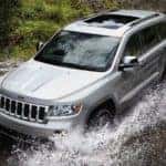 A silver 2013 used Jeep Grand Cherokee is driving through a big puddle splashing up water.