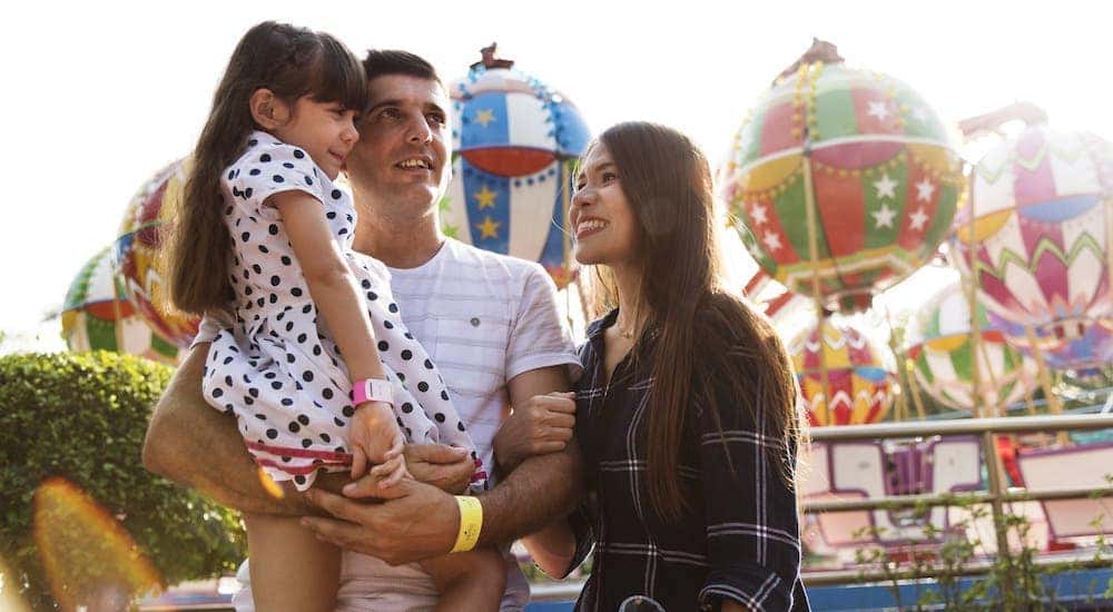 A happy family is at an amusement park in front of colorful rides.