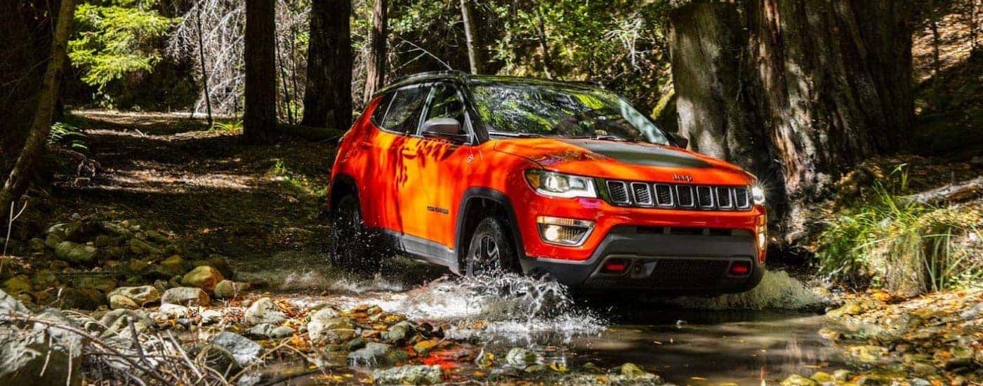 An orange 2019 Jeep Compass Trailhawk is driving through a river in the woods.