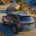 A gray used Jeep Cherokee is driving around a winding road and shown from the rear.