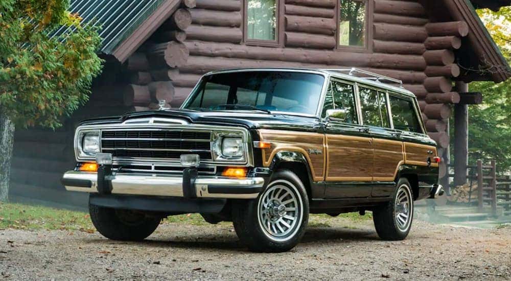 A wood paneled 1991 Jeep Grand Wagoneer is parked in front of a log cabin and would be hard to find at a Jeep dealer in California.