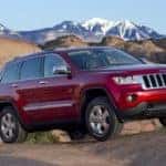 A red 2011 Jeep Grand Cherokee is parked on a hill in front of snow-covered mountains.