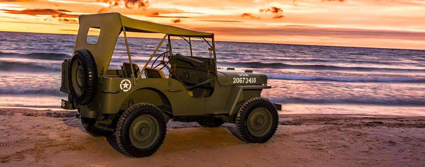 An olive green 1942 Jeep Wrangler Willys is shown from the side while parked on the beach with an orange sunset in the background.