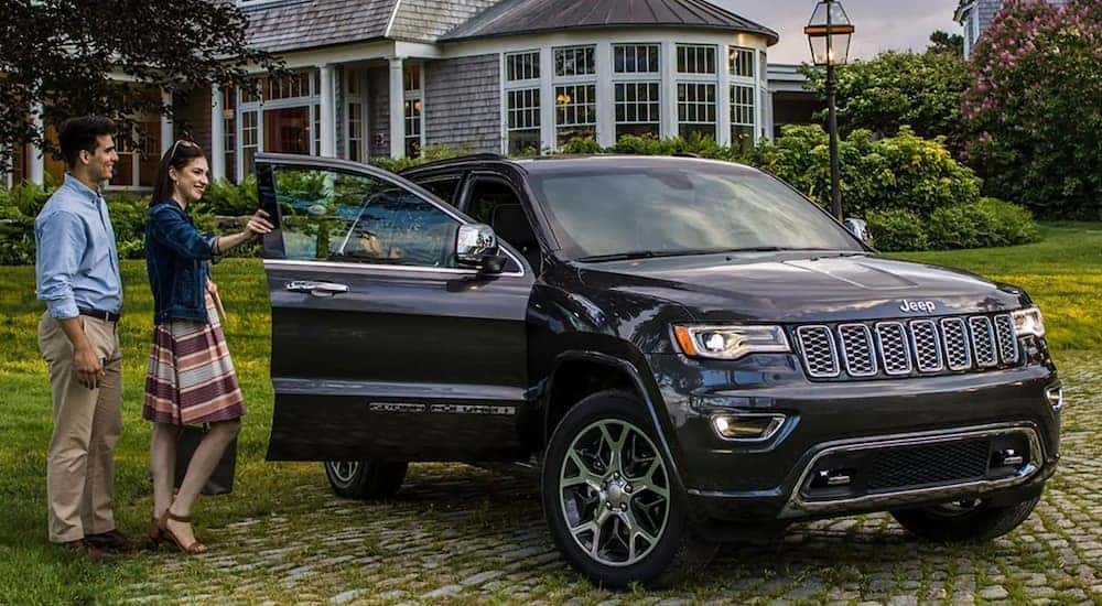 A black 2021 Jeep Grand Cherokee Overland is parked outside of a house as a couple opens the door after reading the Jeep Grand Cherokee Buyers Guide.