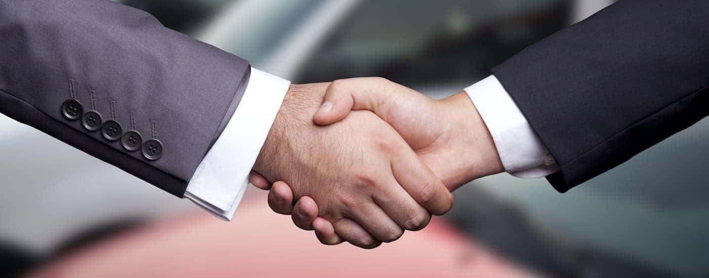 A salesman is shaking the hand of a car buyer at a used car lot.