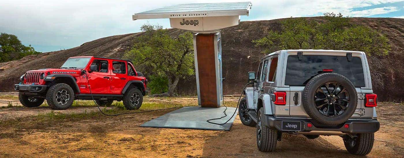 A red and a grey 2021 Jeep Wrangler 4xe are shown at a charging station.