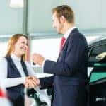 A salesman is shaking hands with a customer at a used car dealer.