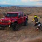 A red 2020 Jeep Gladiator Rubicon is shown from the front at an angle in the desert with a dirt bike passing by.