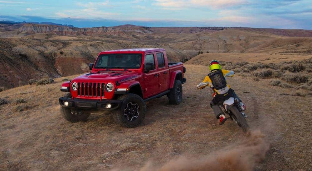A red 2020 Jeep Gladiator Rubicon is shown from the front at an angle in the desert with a dirt bike passing by.