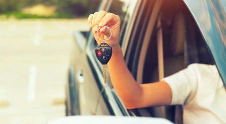 A person is shown holding a car key out the window of a cheap used car for sale.