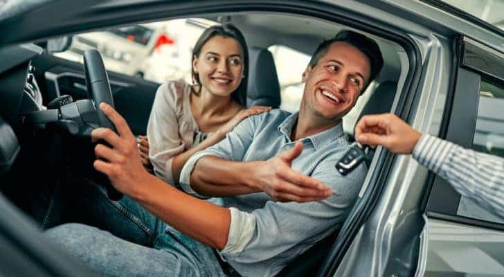 A couple is shown sitting in the front seat of a car as a salesperson is passing a key.